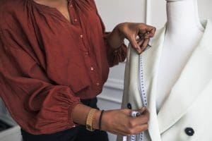 Person in a red blouse measures the lapel of a white jacket on a dress form with a measuring tape.