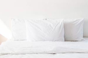 Three white pillows arranged on a white bed with white bedding against a plain white wall.