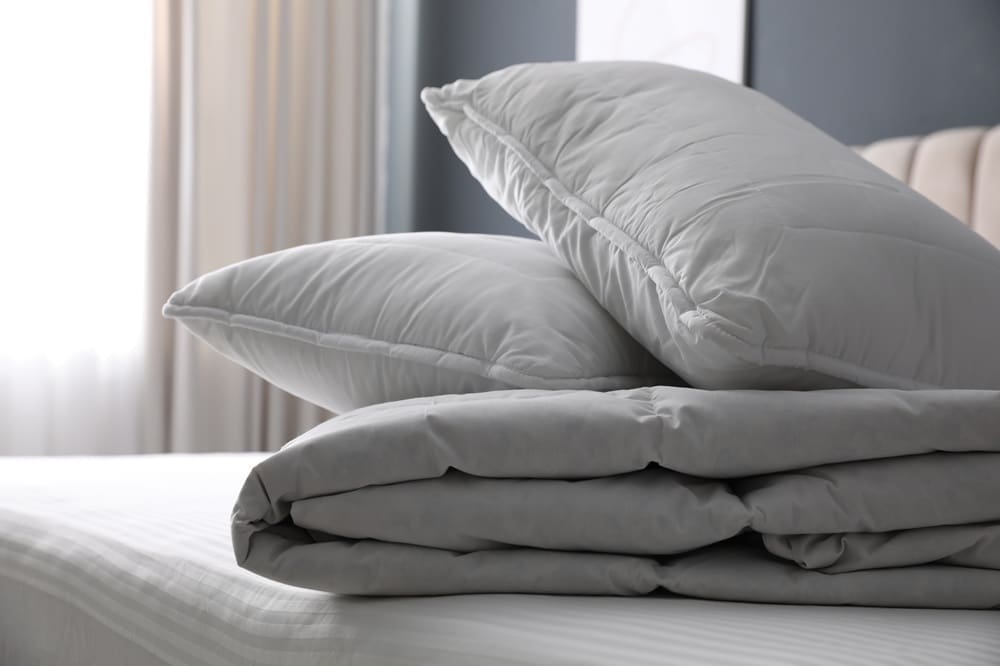 Two gray pillows stacked on top of a folded gray comforter, placed on a neatly made bed in a bedroom with soft natural light.