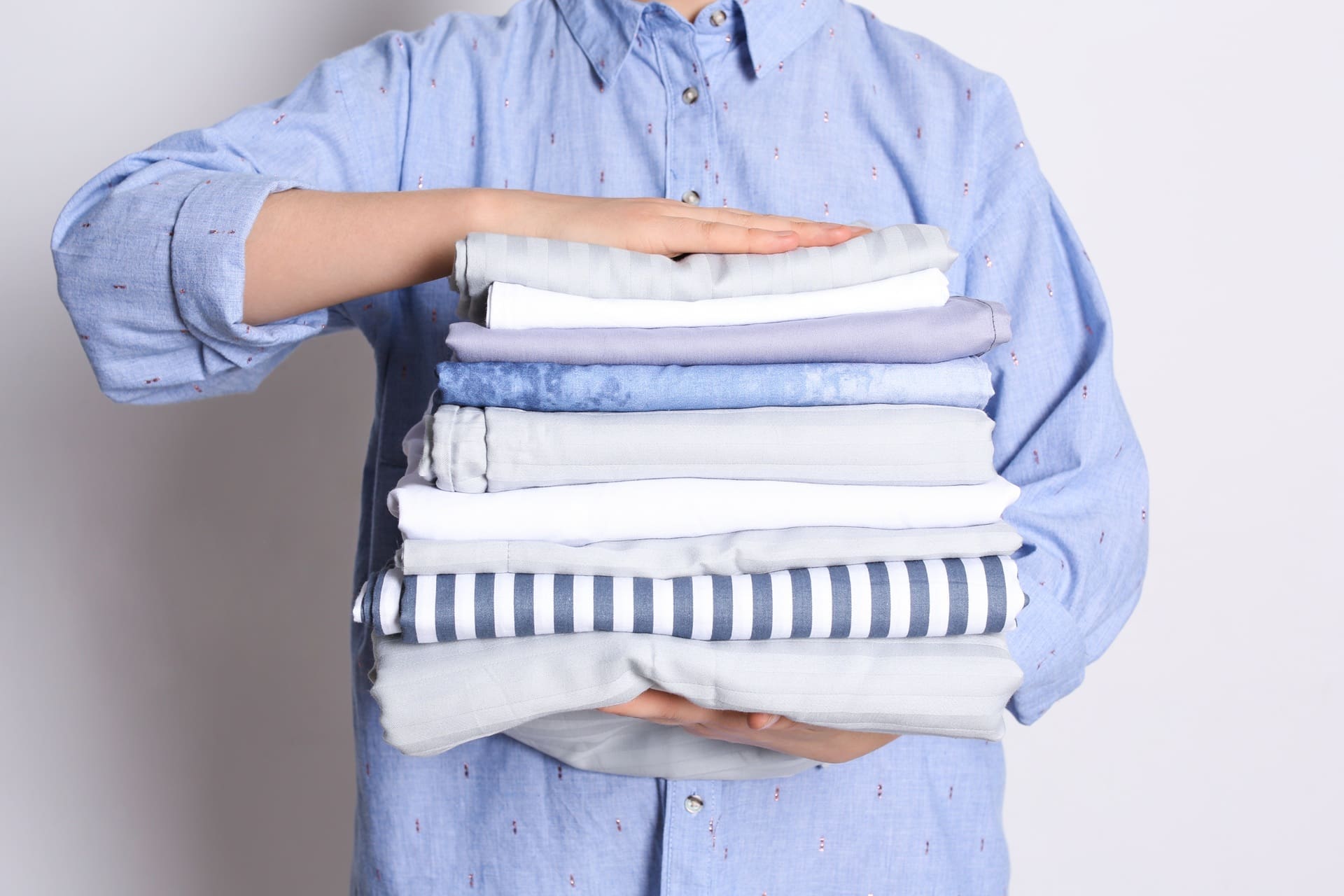A person in a blue shirt holds a neatly folded stack of various white, gray, and blue linens against a plain background.