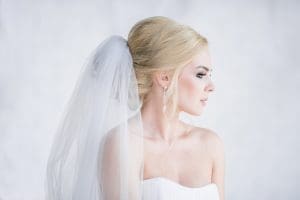 A woman in a strapless white dress with a veil and drop earrings looks to the side against a light background.