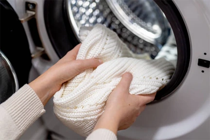 Person placing a white knitted sweater into the drum of a front-loading washing machine.