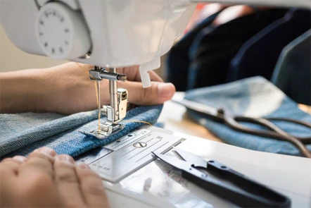 Close-up of hands guiding denim fabric through a sewing machine, with sewing tools and scissors nearby on the table.