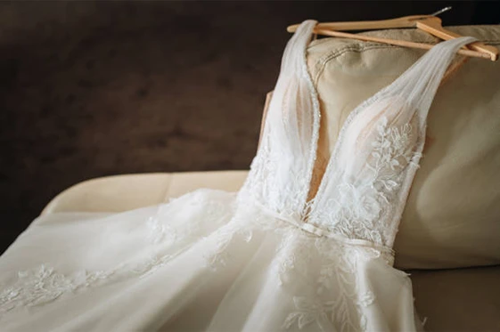A white, sleeveless wedding dress with lace details and a deep V neckline is draped over a beige sofa, displayed on a wooden hanger.