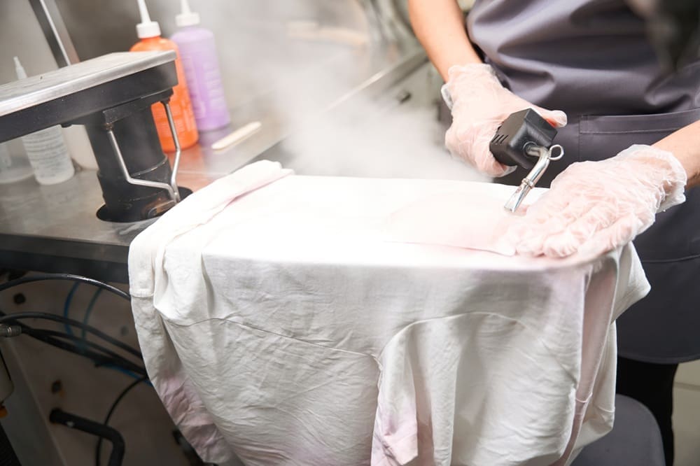 Person using a steam press to iron a white shirt at a dry cleaning facility, with bottles of cleaning products visible in the background.