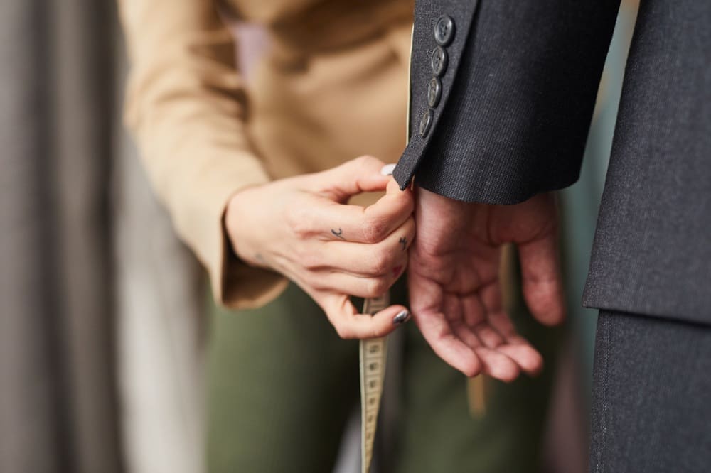 A tailor adjusts the sleeve of a dark suit jacket on a person, holding a measuring tape and checking the fit.