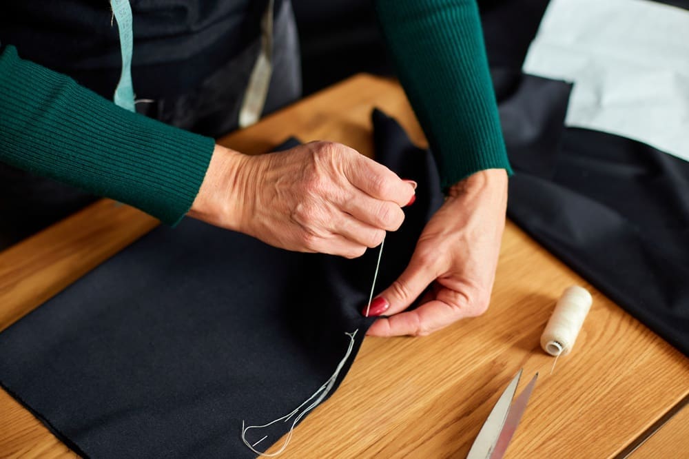 A person hand sewing black fabric with a needle and thread on a wooden table, with scissors and a spool of thread nearby.