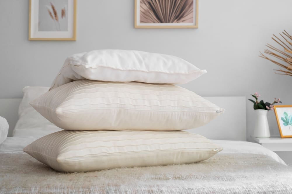 Three pillows stacked on a bed in a neatly decorated bedroom with framed artwork and decorative items in the background.