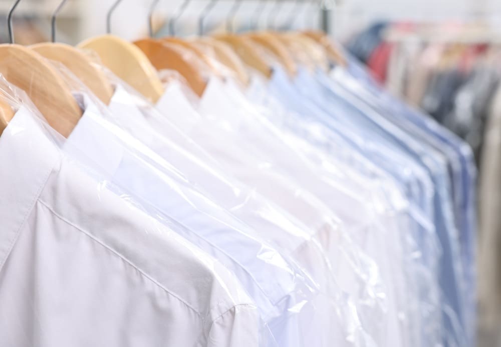 Row of cleaned and pressed shirts covered in plastic garment bags hanging on wooden hangers at a dry cleaner.