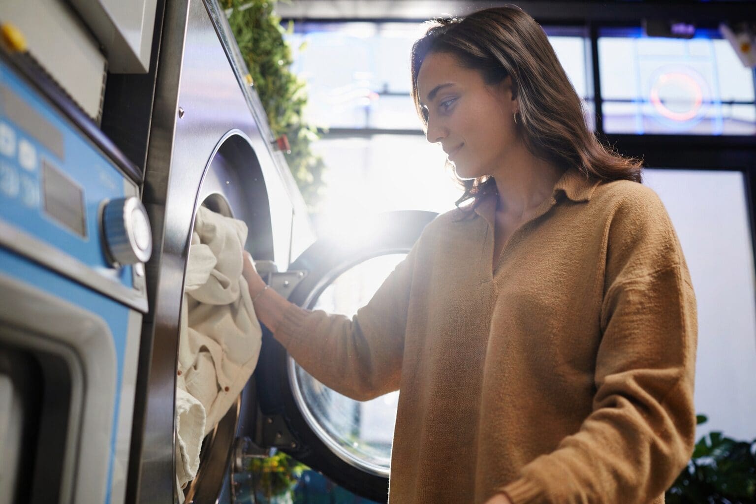A woman wearing a brown shirt loads or unloads laundry from a front-loading washing machine in a brightly lit laundromat.
