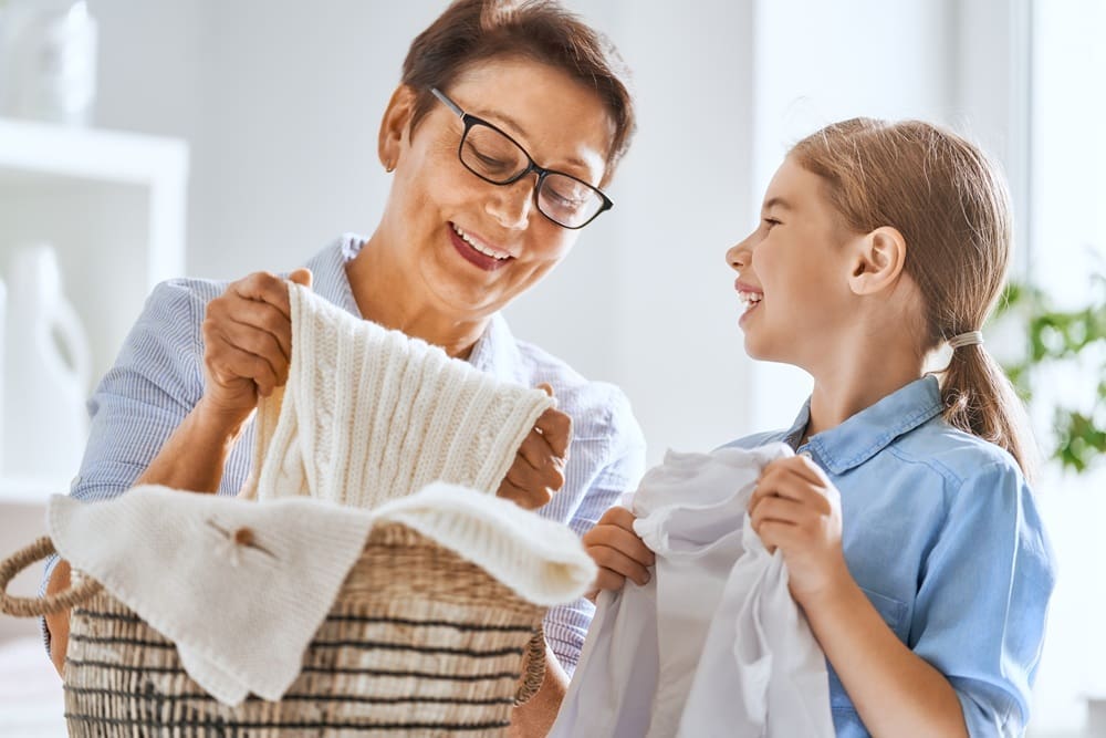 An older woman and a young girl smiling while folding laundry together, holding clothes and a laundry basket in a bright room.