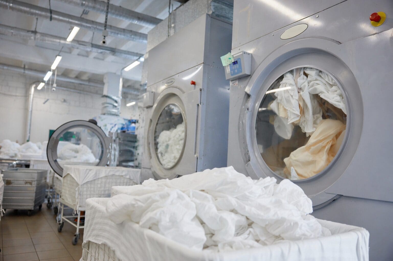 Large industrial washing machines with white linens inside and laundry carts filled with clean linens in a commercial laundry facility.