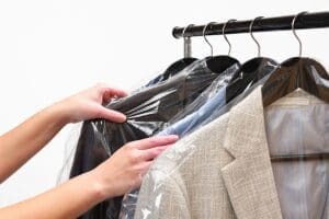 A person’s hands are removing plastic covering from a row of dry-cleaned clothes hanging on a rack.