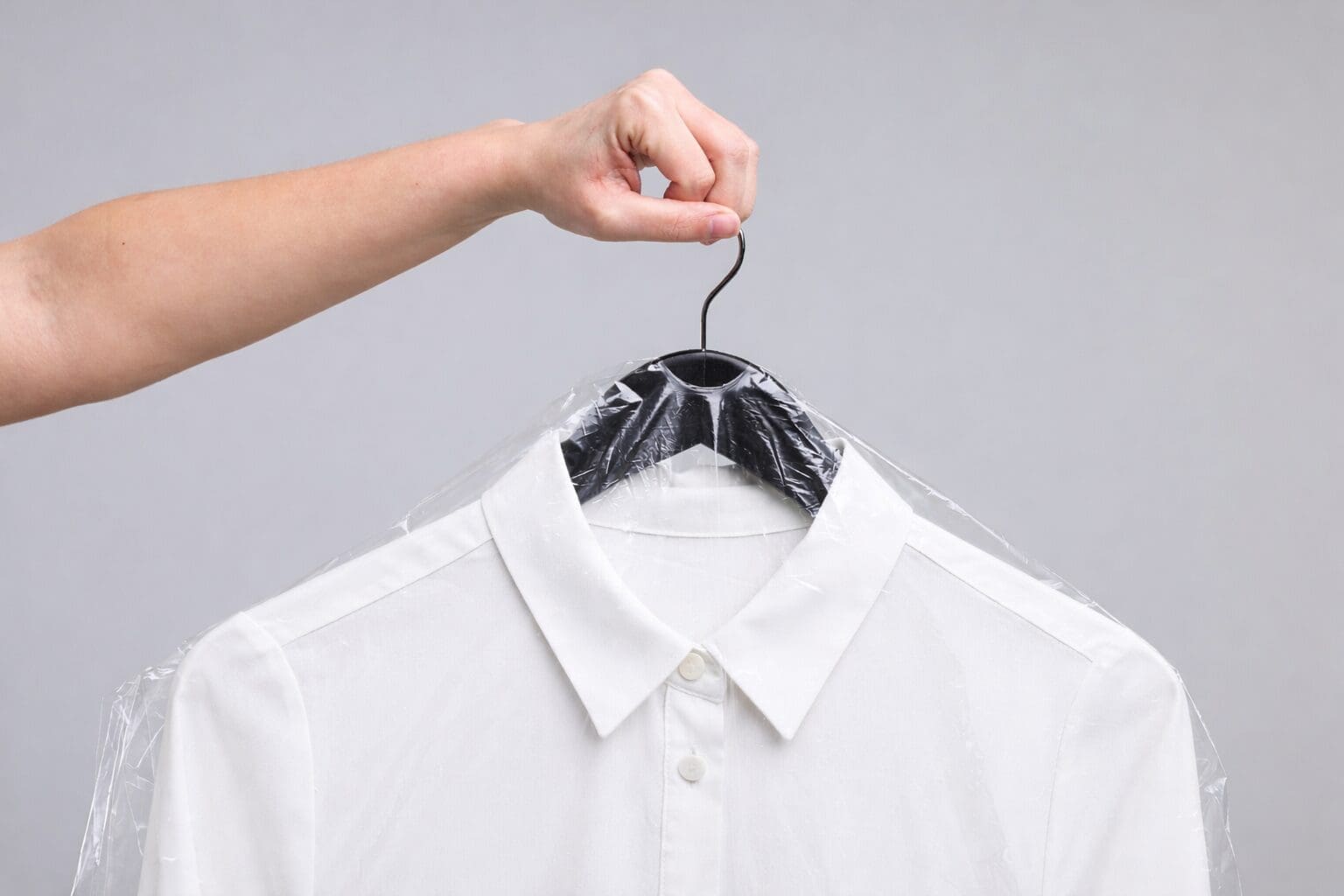 A hand holds a hanger with a white dress shirt covered in clear plastic, typically used for dry cleaning, against a plain gray background.