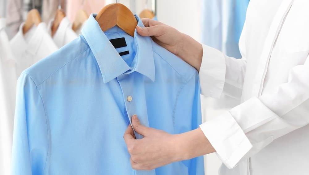 Person holding a light blue dress shirt on a wooden hanger, inspecting its front, with other shirts visible in the background.