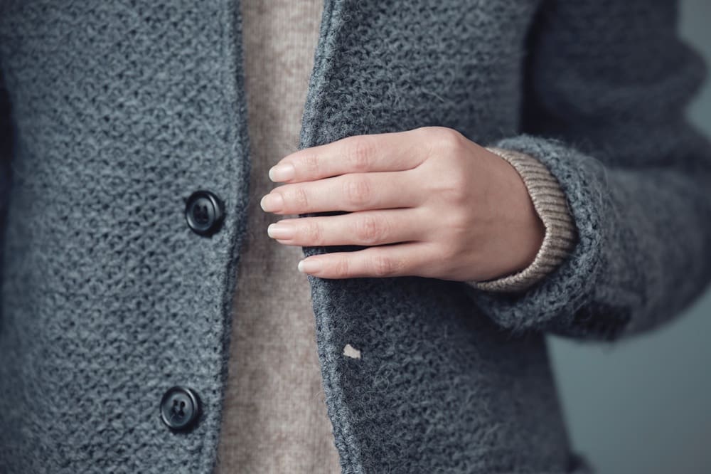 A person wearing a textured gray coat and beige sweater fastens a button on the coat with their right hand.