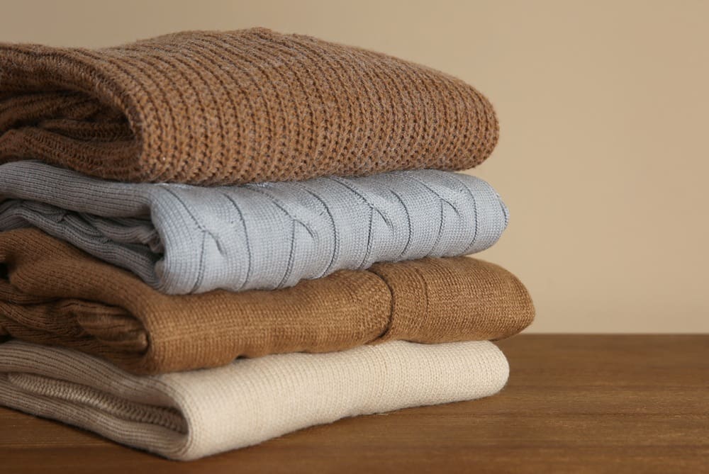 A stack of four folded sweaters in brown, blue, beige, and cream colors sits on a wooden surface against a neutral background.
