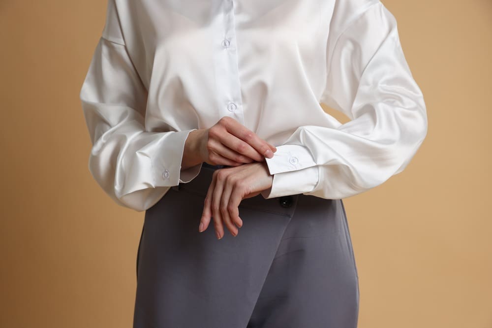 Person in a white button-up shirt and gray pants adjusting a sleeve cuff against a plain beige background. Face is not visible.