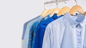 Six shirts in various shades of blue and patterns hang on wooden hangers on a metal clothing rack against a plain light background.