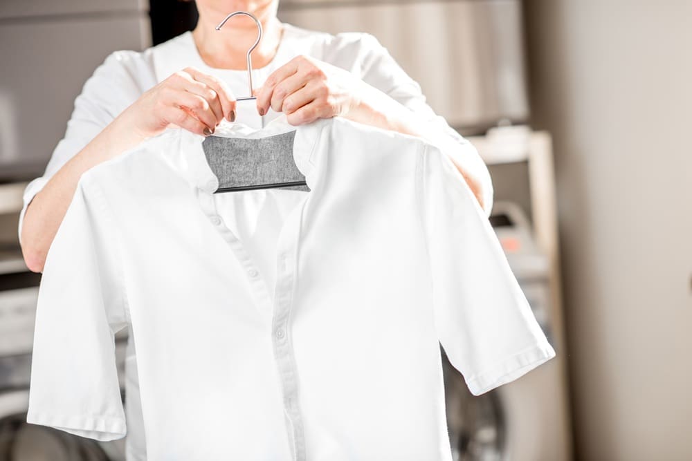 A person holds up a clean white shirt on a hanger, with laundry machines visible in the background.