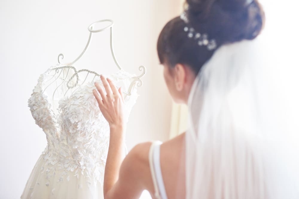 A bride wearing a veil and white dress faces and touches a mannequin displaying a lace wedding gown in a bright room.