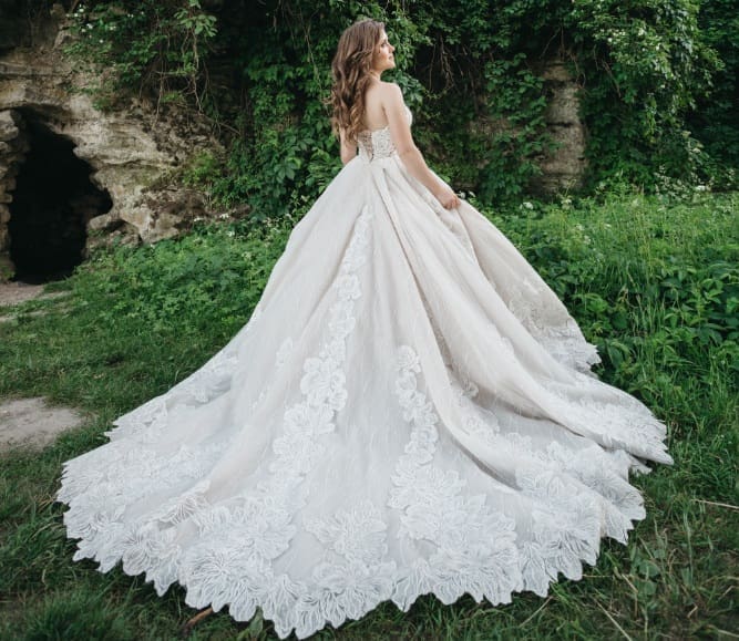 A woman in a white, strapless wedding dress with a long, lace train stands outdoors near greenery and a stone structure.