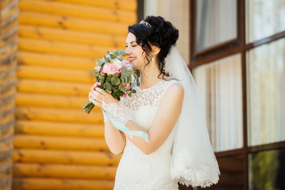 A bride in a white lace dress and veil holds a bouquet of flowers and smiles outside a wooden building.