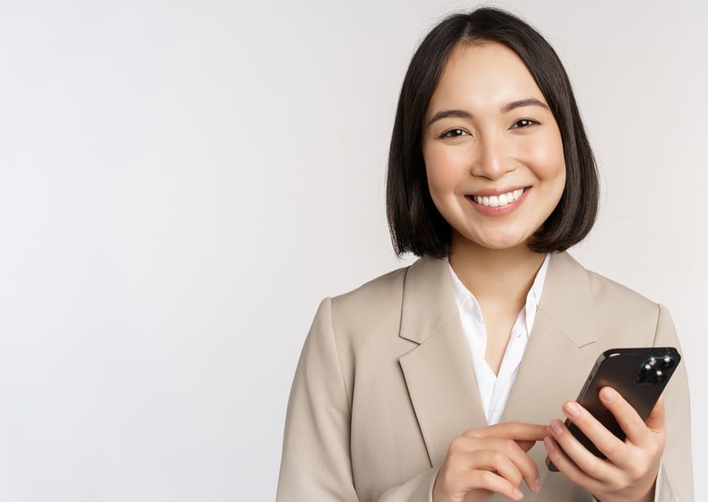 A woman in a beige blazer smiles at the camera while holding and using a smartphone against a plain light background.