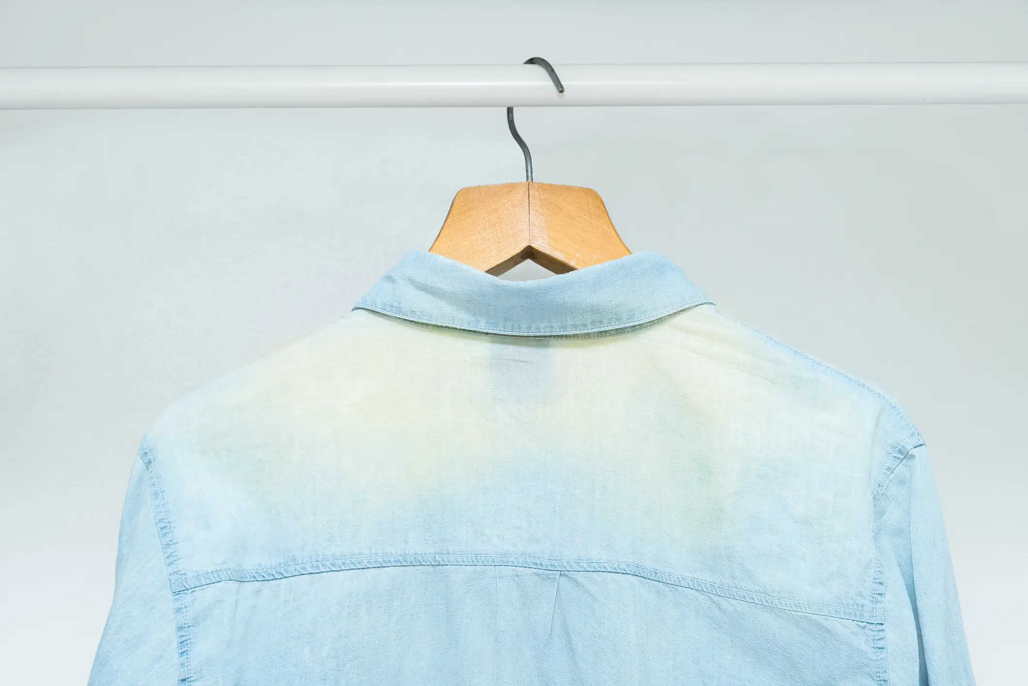 A light blue denim shirt with a yellowish stain on the back, hanging on a wooden hanger from a white metal rod.